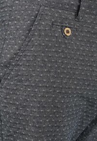 Pantalon gris foncé en tissu texturé, présentant un motif subtil, une fermeture par bouton et des poches latérales.