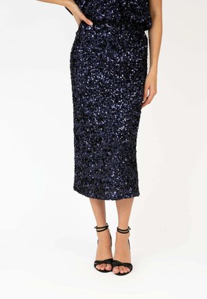 Jupe midi bleu marine à sequins portée avec des talons hauts noirs à bride à la cheville, modèle posant avec une main sur la hanche devant un fond uni.