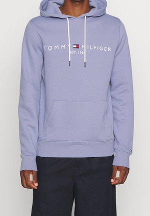 Mężczyzna w lawendowej bluzie z kapturem Tommy Hilfiger z białymi sznurkami i czarnych spodniach, stojący na tle jednolitego jasnego tła.