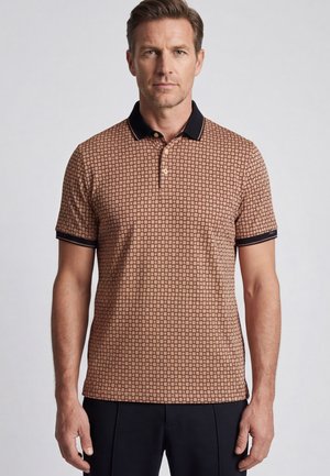 Man draagt een bruin poloshirt met patroon, zwarte kraag en manchetten, staand tegen een effen lichte achtergrond.