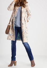 Trinchera con estampado floral en beige, con mangas largas y bolsillos, combinada con una blusa azul clara y jeans oscuros. Zapatos de tacón cuadrado marrones.