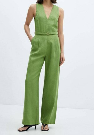 Kvinde iført ærmeløs grøn jumpsuit med dyb V-udskæring, bælte i taljen, vide ben og sorte åbne hæle på ensfarvet baggrund.