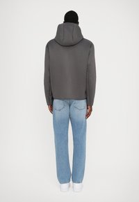 Grå huva jacka med en slät textur, i kombination med ljusblå raka jeans och vita sneakers, sedd från baksidan.