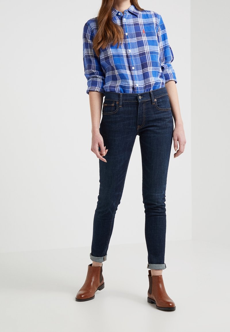 Femme portant une chemise en flanelle bleue, un jean skinny foncé retroussé et des bottines marron, debout devant un fond uni.