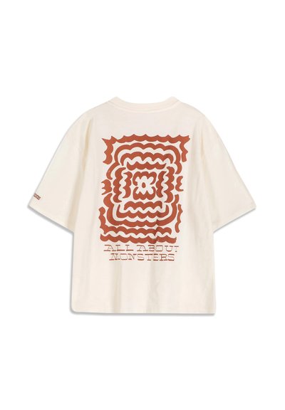 T-shirt crème à manches courtes avec motif carré ondulé de couleur rouille et texte "ALL ABOUT MONSTERS" dans le dos.
