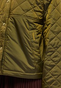 Pihepuha olívazöld dzseki patentgombokkal, oldalzsebbel, amelyen arany logó található, és sima, könnyű textúrájú.