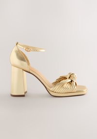 Zlaté sandále na vysokom podpätku s masívnym blokovým podpätkom, uzlový dizajn vpredu, syntetický materiál a nastaviteľný remienok okolo členka.
