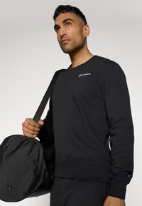Sweatshirt noir à manches longues, col rond, avec des poignets et un ourlet côtelés. Présente un logo brodé sur la poitrine. Le modèle porte un sac de sport noir.