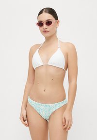 Biely rebrovaný bikiny top s trojuholníkovými košíčkami, spárovaný so svetlomodrými spodnými dielmi s kvetinovým vzorom. Modelka nosí ružové oválne slnečné okuliare.