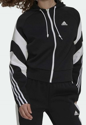 Czarno-biała bluza z kapturem i zamkiem Adidas oraz pasujące spodnie z białymi paskami na rękawach i nogawkach, noszone przez osobę z kręconymi włosami.