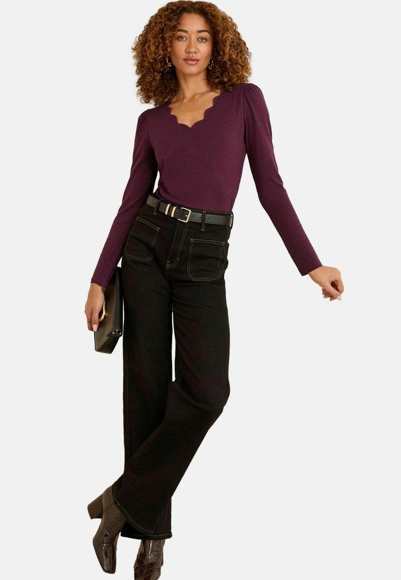 Göndör hajú nő fitted lila felsőben, amelynek fodros dekoltázsa van, fekete, oversize nadrágban, fekete övvel és barna sarkú csizmában, clutch-ot tartva.