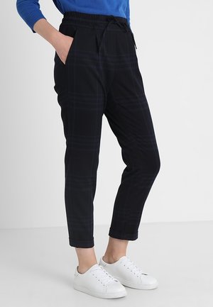 Person trägt schwarze karierte, schmal geschnittene Hose mit elastischem Bund und weißen Sneakers, steht vor einem schlichten Hintergrund.