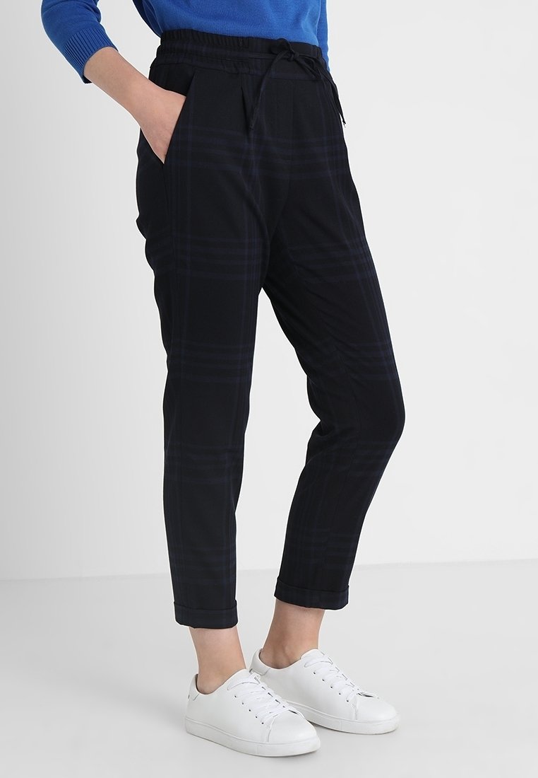Personne portant un pantalon noir à carreaux fuselé avec une taille élastique et des baskets blanches, debout devant un fond uni.