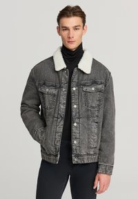 Giovane uomo che indossa una giacca di denim grigia lavata con colletto in pelliccia bianca sopra un dolcevita nero e pantaloni neri, in piedi contro uno sfondo neutro.