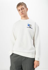 Giovane uomo con capelli biondi corti, che indossa una felpa bianca con un logo blu e pantaloni grigi scuri, in piedi su uno sfondo bianco.