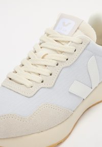 Sneaker azzurri chiari con materiale текстurizzato, dettagli in suede beige, logo bianco sulla linguetta e lacci color crema. Suola in gomma.