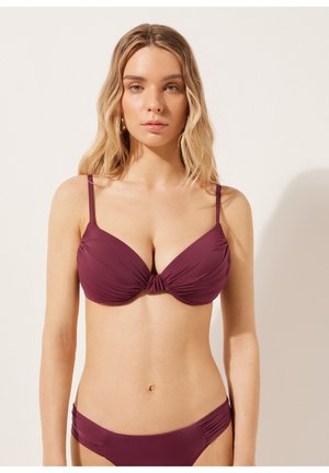 Frau mit langen blonden Haaren, die einen dunkelroten Bikini mit gerafften Stoffdetails trägt und vor einem schlichten hellen Hintergrund steht.