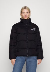 Svart pufferjacka med hög krage, zipfront och horisontell quiltning. Har broderad logotyp på vänster bröst och sidofickor.