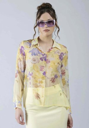 Donna che indossa una camicetta trasparente a fiori giallo chiaro, gonna gialla abbinata e grandi occhiali da sole viola, in piedi davanti a uno sfondo semplice.