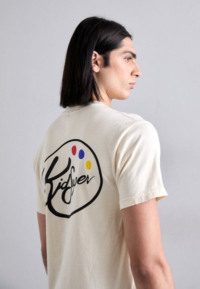 Beige cotton t-shirt with short sleeves, featuring a black logo and colorful paint dots on the back. Simple, casual fit.