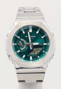 Sølv G-Shock-klokke med åttekantet kasse, grønn skive og digital skjerm. Sølvfarget metalllenke med teksturerte detaljer og justeringsknapper.