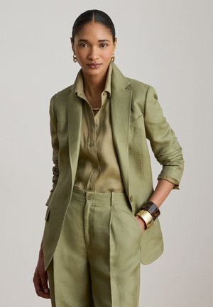 Kvinde iført olivengrøn blazer, matchende bukser og skjorte med guldringeøreringe og armbånd i blandede metaller, poserer med hånden i lommen.