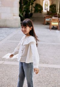 Chemise blanche texturée à boutons avec col à plusieurs couches, associée à un jean bleu clair à jambes larges. Cadre extérieur avec un éclairage doux et naturel.
