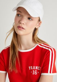 Rode t-shirt met korte mouwen en witte rand, met de opdruk "FRANKRIJK 16". Draagt een witte pet en ringoorbellen. Glad materiaal, atletisch ontwerp.