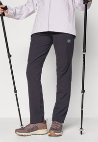 Hiking pants in dark gray, featuring a zippered pocket, stretchy fabric, and a logo detail. Paired with brown and gray hiking shoes.