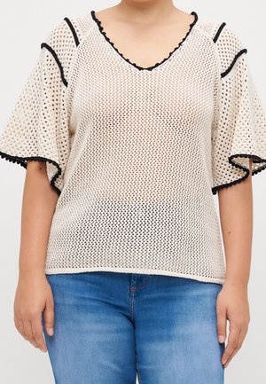 Woman wearing a beige open-knit short-sleeve top with black trim and blue jeans, standing against a plain background.