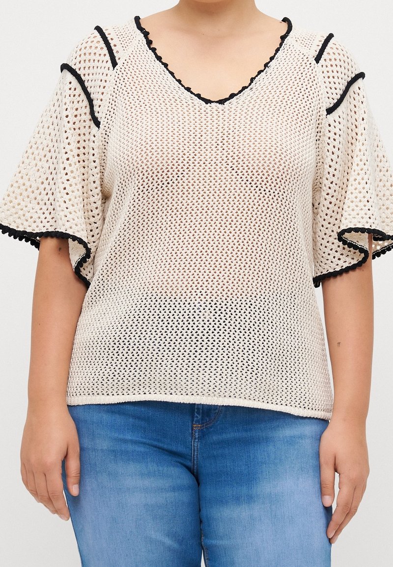 Femme portant un haut à manches courtes en maille beige avec bordure noire et un jean bleu, debout devant un fond uni.