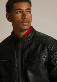 Czarna skórzana kurtka z kołnierzem, zapinana na zamek z przodu, oraz dwiema zapinanymi na zamek kieszeniami na klatce piersiowej. Posiada pikowane akcenty na ramionach i gładką fakturę.