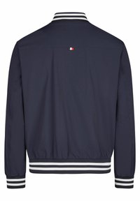 Marineblauwe jas met een gladde textuur, gestreepte witte en zwarte ribboorden aan de mouwen en kraag, met een klein driekleurig logo op de achterkant.