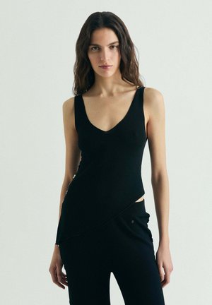 Mujer con cabello largo y oscuro, vistiendo una blusa negra sin mangas con botones en el costado y pantalones negros a juego, sobre un fondo liso.