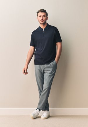 Man staat tegen een beige muur, draagt een zwart shirt met korte mouwen en een ritskraag, grijze broek, witte sneakers, met één hand in zijn zak.