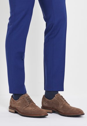 Brązowe brogsy z zamszu noszone z granatowymi skarpetami i dopasowanymi niebieskimi spodniami na jednolitym jasnym tle.