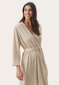 Beige Wickelkleid mit langen Ärmeln, das eine vertikale Rippstruktur und einen Stoffgürtel in der Taille aufweist. Leicht taillierte Silhouette.