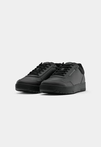 Paire de baskets basses noires avec lacets, estampillées "Tommy Hilfiger" sur le côté et la languette, sur un fond clair uni.