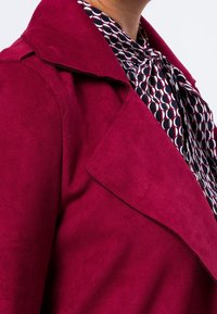 Fuchsia suède jas met een brede kraag, met daaronder een gebloemde blouse met veelkleurige cirkels en een gestrikt neklint.