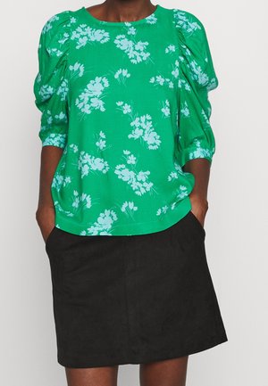 Kvinde iført en grøn floral bluse med puffede ærmer, stoppet ned i et sort knælængde skørt, hænderne i skørtets lommer, foran en ensfarvet baggrund.