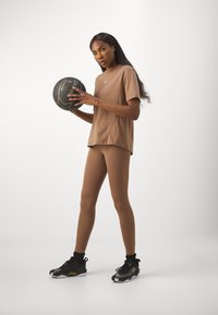 Mujer con ropa de deporte marrón sosteniendo un balón de baloncesto negro, de pie contra un fondo claro, usando zapatillas negras y doradas.