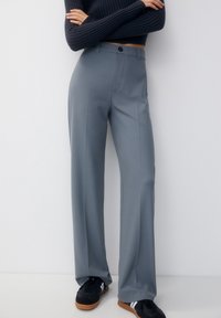 Grijze, hooggerimpelde broek met een rechte pijp, platte voorkant en een knoopsluiting. Gemonteerd met zwarte sneakers.