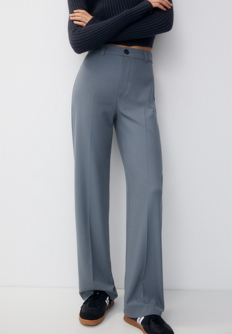 Grijze, hooggerimpelde broek met een rechte pijp, platte voorkant en een knoopsluiting. Gemonteerd met zwarte sneakers.