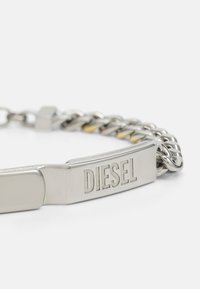 Pulseira de metal prateado com um design de corrente grossa, apresentando uma placa gravada com "DIESEL", acabamento suave e pontos polidos.