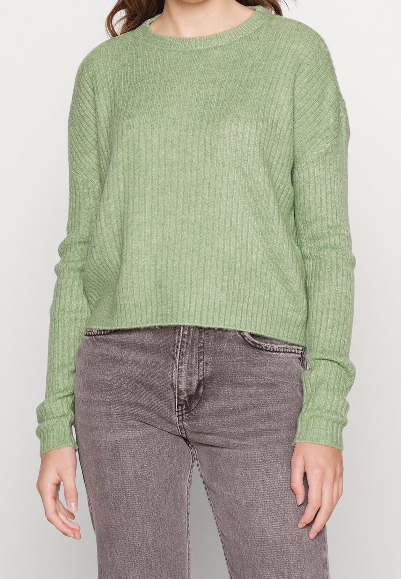 Femme portant un pull à manches longues côtelé vert clair associé à un jean gris délavé taille haute, debout devant un fond neutre.