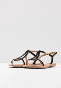 Black suede sandals feature crisscross straps, a beige sole, and gold hardware. Open design with a flat profile and minimalistic accents.