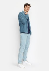 Camicia di velluto a coste celeste sopra un top blu navy, abbinata a jeans chiari e sneakers bianche. Il modello è in profilo su uno sfondo neutro.