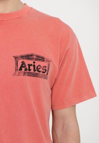 Koraliowa bawełniana koszulka w luźnym kroju, z czarnym nadrukiem przedstawiającym klasyczną strukturę oraz napisem "Aries" na piersi.