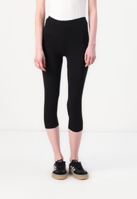 Svart, avkortede leggings med en glatt tekstur og elastisk midje, kombinert med svarte joggesko med hvite og grå striper.