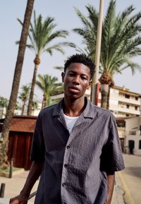 Jeune homme portant une chemise texturée gris foncé à boutons, debout à l'extérieur près de palmiers et de bâtiments par une journée ensoleillée.